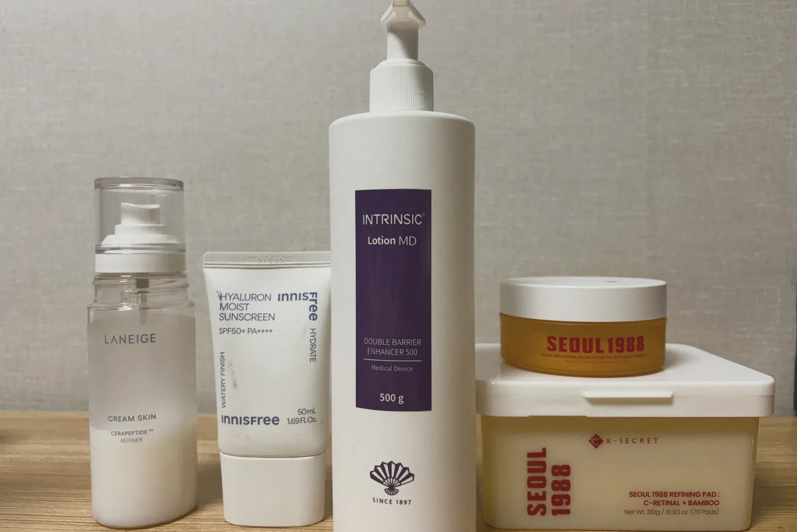 A frosted mist bottle, a white sunscreen tube, a tall white pump bottle with a purple label, and a tub of pads topped with an orange jar, lined up on a wooden shelf against pale wallpaper.