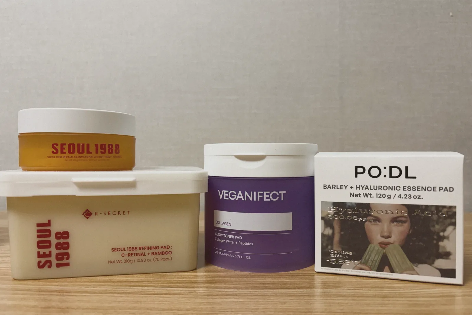 An orange jar of eye patches stacked on a tub of retinal pads, beside a purple jar of toner pads and a white box of essence pads, on a wooden shelf.